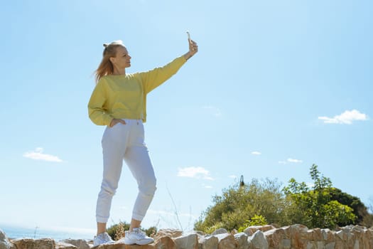 A girl of European appearance is standing in white pants and a yellow jacket taking a selfie overlooking the sea. High quality photo