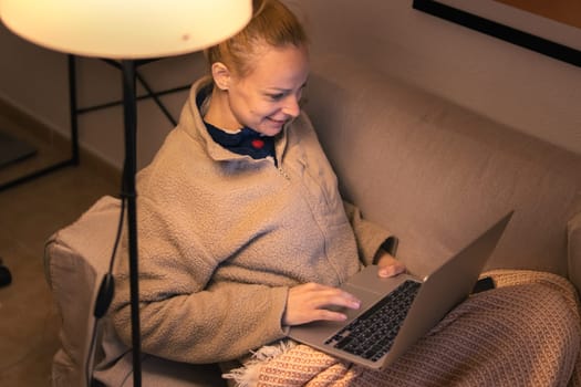 a woman of European appearance sits in the living room on the sofa covered with a brown blanket, works on a laptop, there is a place for an inscription. High quality photo