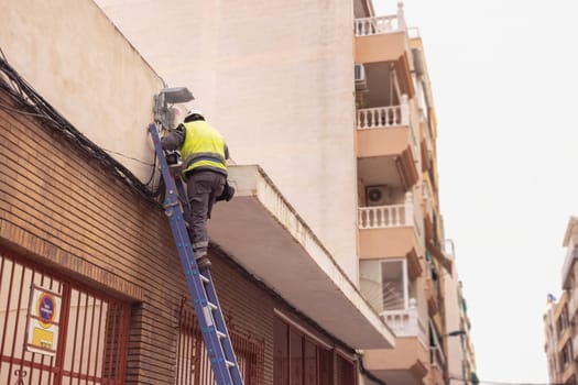A man in a reflective vest stands on a stepladder doing electrics. A man in work clothes repairs the cable at home. High quality photo