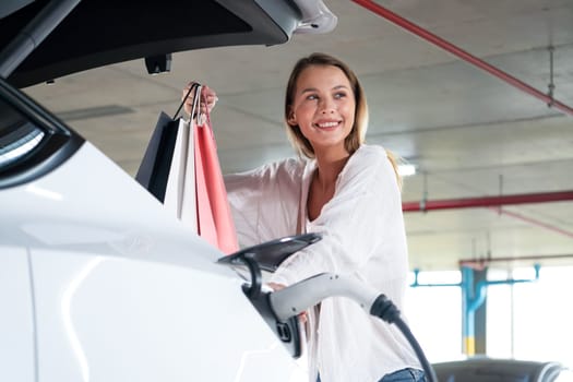 Young woman travel with EV electric car to shopping center parking lot charging in downtown city showing urban sustainability lifestyle by green clean rechargeable energy of electric vehicle innards