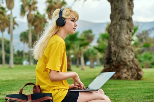 Teenage student sitting in park with laptop. Female teenager in headphones with backpack writing in notebook. Education, adolescence, high school, college, youth concept