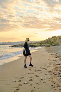 Young teenage female on beach in morning. Hipster teenager in headphones walking along seashore with picturesque colorful sea nature. Summer sunrise, freedom vacation beauty youth concept