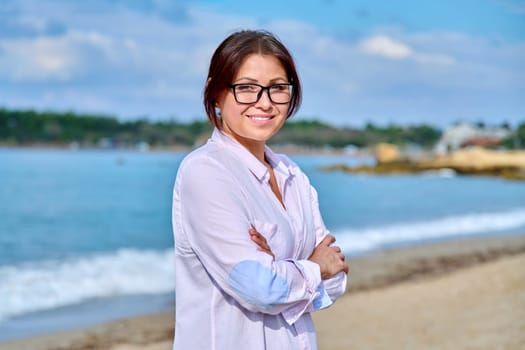 Outdoor portrait of mature smiling woman on sea vacation. Confident successful middle aged female with folded hands looking at camera. Leisure, vacation, lifestyle, people 40s age concept
