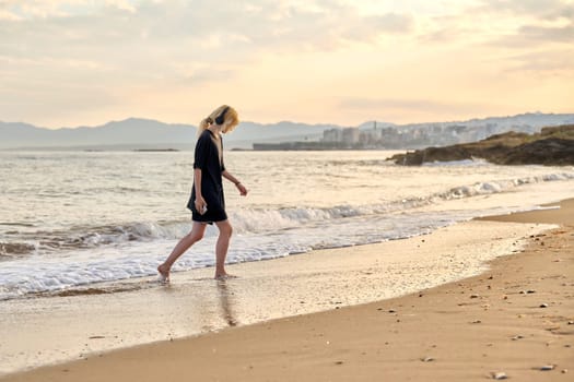 Young teenage female on beach in morning. Hipster teenager in headphones walking along seashore with picturesque colorful sea nature. Summer sunrise, freedom vacation beauty youth concept