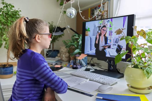 Girl child sitting at home at table with computer listening online lesson. Woman teacher on computer screen teaching remotely individually to schoolgirl student. E-education, virtual distance learning