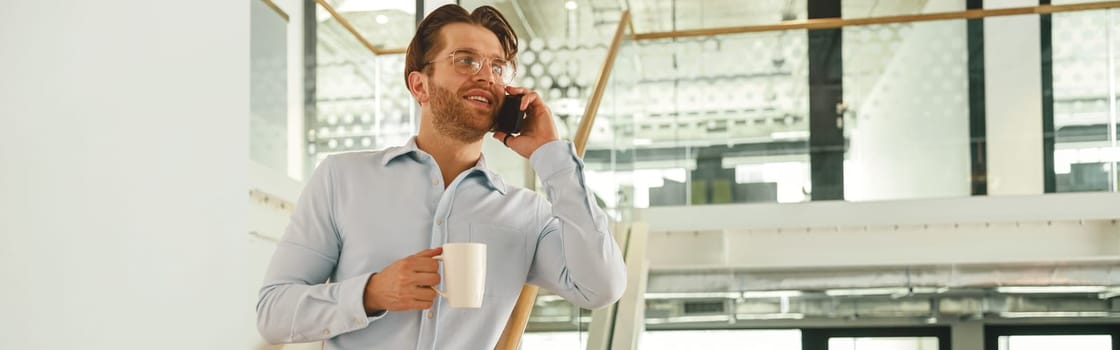 Male sales manager with coffee cup talking phone with client while standing in coworking on stairs