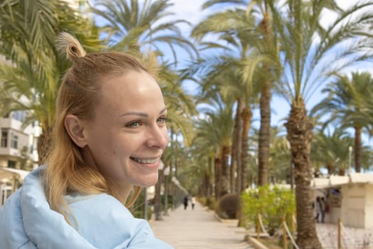 a girl of European appearance with blond hair, in a blue jacket, stands on the embankment, smiles around green palm trees. A beautiful seascape. High quality photo