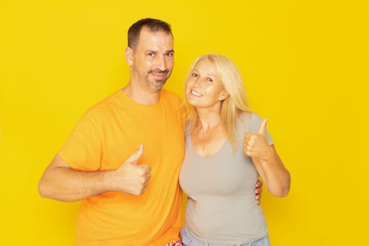 Happy caucasian couple in their 40s showing thumbs up and looking at camera isolated on yellow background