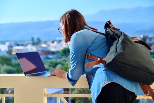 Confident serious mature businesswoman with laptop outdoors. Middle-aged female in denim shirt headphones glasses with backpack looking at laptop screen. Work, business, technology, people 40s age