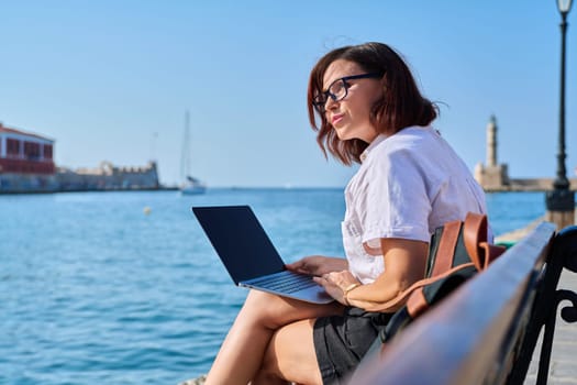 Business mature woman sitting outdoor using laptop. Female 40s with backpack sitting on bench on sea promenade working with laptop. Summer, business travel, freelancing, vacation, people concept