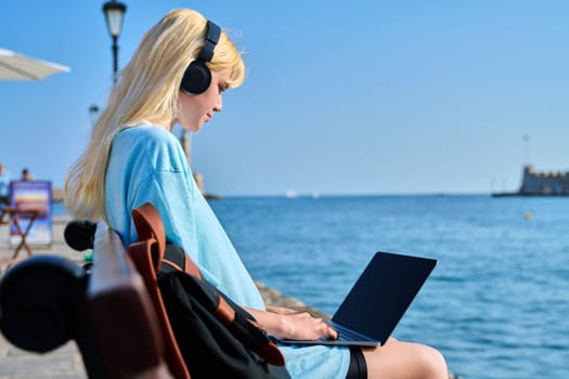 Young woman relaxing in headphones with a laptop on the seashore, in a touristic summer city. Beautiful teenage blonde enjoying her vacation. Youth, vacation, technology, tourism concept