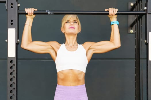 Strong fit muscular female in activewear doing pull ups on metal Horizontal bar while performing workout at fitness club against gray background looking up