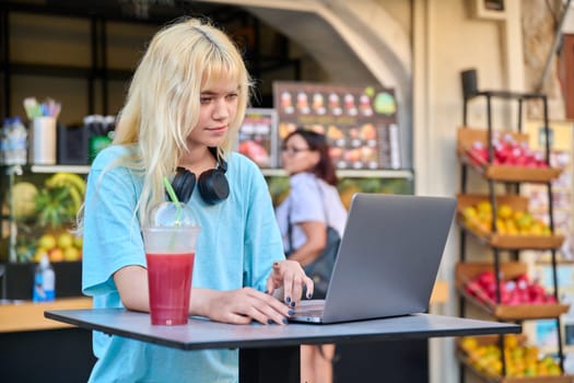 Summer in the city, young teenage woman in outdoor fruit juice bar with fruit fresh juice using laptop. Lifestyle, youth, healthy food drinks, leisure concept
