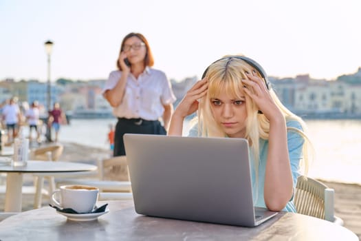 Surprised teenage female with headphones looking at a laptop in an outdoor cafe. Beautiful emotional young blonde on a summer day at a table with a cup of coffee on the seaside promenade.