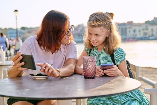 Mother and daughter child together in an outdoor cafe on the waterfront. Family, mom and preteen girl using smartphones, with coffee, drinks. Lifestyle, family, parent and child concept