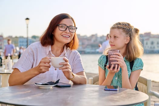 Happy mom and child daughter are resting together. Mother and preteen girl talking laughing in outdoor cafe, seaside city embankment background. Lifestyle, family, friendship, parent and kid concept