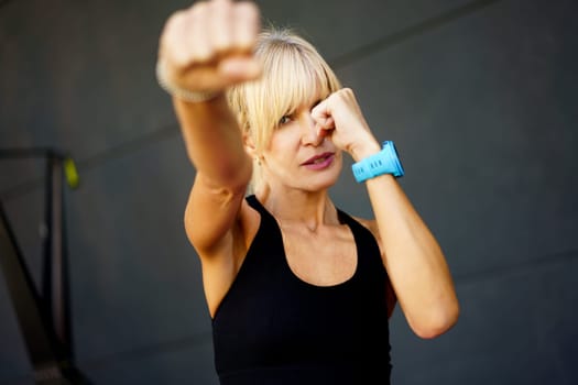 Confident young fit blond haired female wearing black tank top performing martial art punch in fitness gym against gray background looking at camera