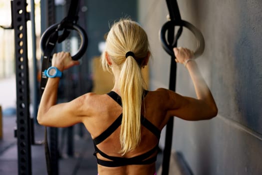 Back view of strong muscular female exercising with fitness gymnastic ring in modern gym club during functional workout against blurred background