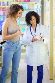 Adult female pharmacist in white robe, looking down while standing with side view of smiling female customer near shelves with assorted medical supplies and explaining medication details in pharmacy