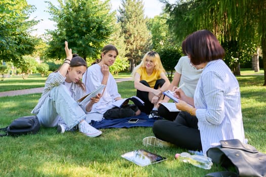 Outdoor, group of students with female teacher. Teenagers and mentor teacher talking sitting on grass in college park. Back to school, back to college, high school, education, teenagers concept