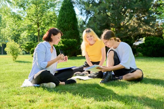 School teacher, psychologist, social worker talking to teenagers. Woman with teenage schoolchildren sitting on grass on campus lawn. Education, high school, psychology, mental health, adolescence