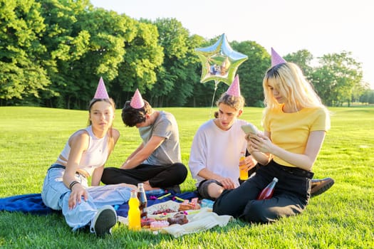 Birthday party outdoor, picnic. Group of teenagers in festive hats celebrating birthday, sitting on the grass, laughing having fun, summer sunny day