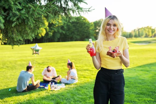 Beautiful teenage girl in festiv hat on birthday with cake and candles. Happy blonde female at outdoor picnic party holding cake with candles 17. Adolescence youth age beauty, holiday birthday concept