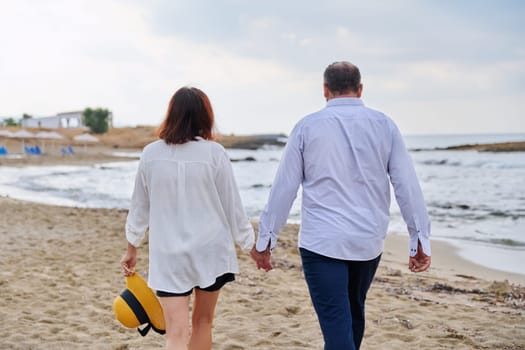 Happy middle aged couple walking together on beach, back view. Mature man and woman holding hands walking along seashore. Relationship, lifestyle, vacation, tourism, sea nature, people concept