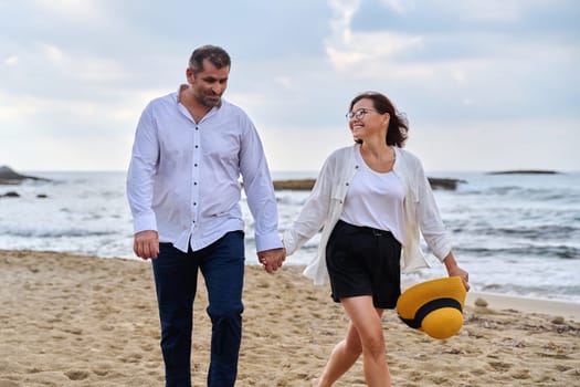 Happy middle-aged couple walking together on the beach. Mature man and woman holding hands walking along seashore. Relationship, lifestyle, vacation, tourism, sea nature, people concept