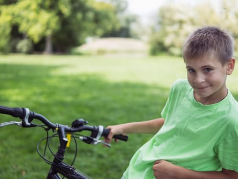 a teenager boy of European appearance with short blond hair in a green T-shirt stands with a bicycle in the park.looks at the camera on the left there is a place for an inscription. High quality photo