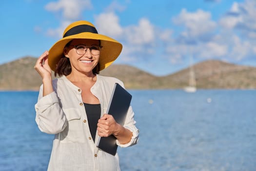 Portrait of happy mature woman in straw hat with laptop in hand on beach, looking at camera, copy space. Freelance, business, remote work, leisure, relaxation, tourism, technology, middle aged people