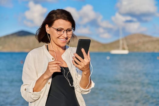 Middle-aged smiling woman relaxing on beach wearing headphones with smartphone. Mature female listening audio podcast, watching video. Leisure technology summer vacations, seaside resorts, people 40s