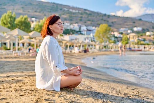 Mature woman sitting in lotus position meditating on the beach. The female enjoys sea nature, the setting sun, summer vacations. Lifestyle, tourism, travel, beauty, health, mature people concept