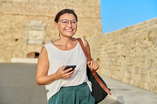 Outdoor portrait of smiling middle-aged woman walking with smartphone in backpack through an old tourist town. 40s age, beauty, health, active lifestyle, travel tourism concept