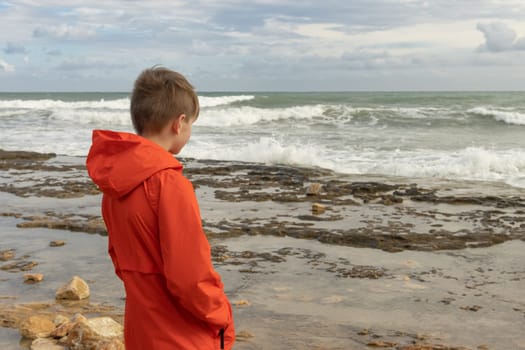 a teenager in an orange windbreaker stands on the beach with his back to the camera and looks at the sea, there is a place for an inscription. Seascape. High quality photo