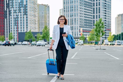 Mature confident business woman walking with suitcase, city background. Smiling middle agged woman with smartphone luggage, looking at camera. Business trip, travel, city, work, lifestyle, people 40s