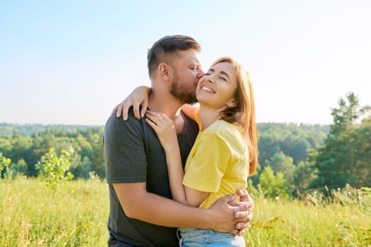 Outdoor portrait of happy middle age couple in love hugging kissing, background summer nature of wild meadow. People 40s, relationships, holidays, lifestyle concept