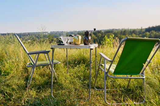 Set of folding furniture for camping, table and chair, cups utensils for brewing fresh coffee, summer nature of wild meadow, sunset on horizon. Summer, holidays, weekends, lifestyle, camping concept