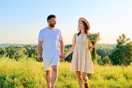 Happy middle age couple holding hands, summer nature wild meadow. Smiling man and woman walking together, with bouquet of wild flowers. People 40s, family, relationships, leisure, holidays, lifestyle