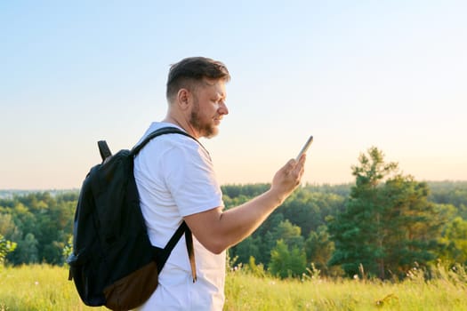 Middle aged bearded man with smartphone backpack on hike, making video call, talking looking at webcam. Wild summer nature meadow forest background. Leisure, technology, adventure, travel, people