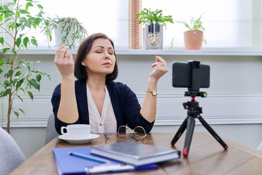 Business mature woman taking break, meditating at home workplace in between online work. Relaxation, stress relief, harmony, balance concept