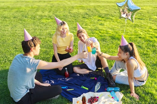 Teenage birthday party picnic on grass in park. Group of teenagers having fun, congratulating girl on her 17th birthday, drinking colored fruit and soft drinks. Age, adolescence, fun, holiday concept