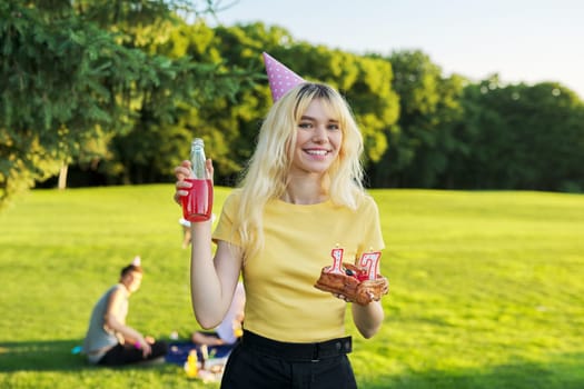 Beautiful teenage girl in festiv hat on birthday with cake and candles. Happy blonde female at outdoor picnic party holding cake with candles 17. Adolescence youth age beauty, holiday birthday concept