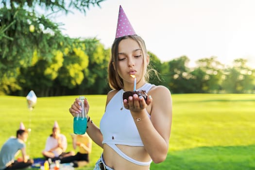Birthday, teenage girl in festiv hat with cake blowing out candle at outdoor party. Picnic in nature, happy having fun female posing looking at camera. Adolescence, holiday, birthday, celebration, age