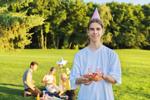 Happy guy teenager in birthday hat with cake with candles 17. Outdoor picnic party with friends in park on grass. Adolescence, having fun, holiday, celebration, birthday, age concept