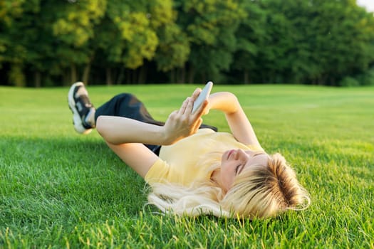 Young teenage woman lying on green grass using smartphone, blonde female resting in park on lawn, in summer sunset light