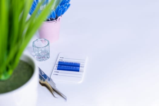 Materials for eyelash extensions.two tweezers,artificial blue eyelashes on a white background. in the background is a pink bucket with brushes for eye. On the side there is a place for an inscription