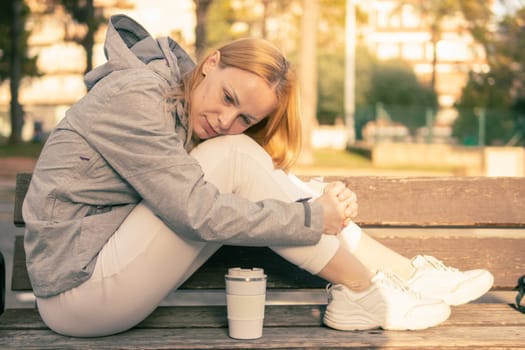 a girl with blond hair sits on a bench with her feet and lowered her head to her feet, the girl sits in a gray jacket and white sweatpants, there is a white thermo mug nearby. High quality photo