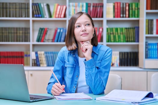 Portrait of smiling young female university student sitting at desk with laptop in library classroom of educational building. Knowledge, higher education, youth concept