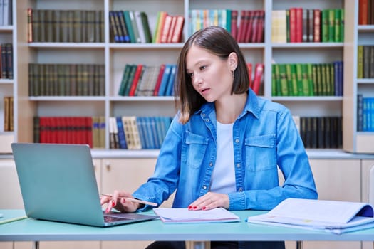 Young female university student studying inside library, typing on laptop computer. Knowledge, education, youth, college university concept